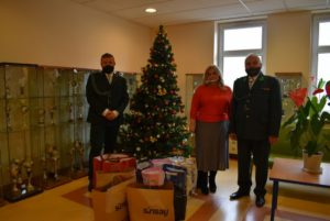 Dwóch mężczyzn w mundurach myśliwskich i kobieta przy choince, przed nimi paczki z prezentami.