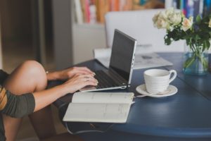 Dłonie na klawiaturze laptopa, obok na stoliku notes i biała filiżanka.