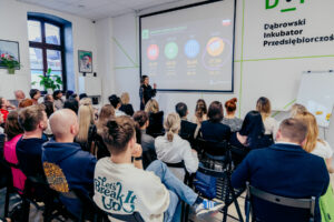 Grupa osób na szkoleniu, które odbywa się w jasnym pomieszczeniu. Prowadząca stoi przed ekranem, na którym wyświetlana jest prezentacja z danymi liczbowymi.