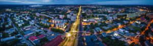 Panorama miasta, oświetlona główna arteria, po jej stronach m.in. bloki mieszkalne oraz mniejsze i niższe budynki. Na dalszym planie drzewa i zieleń.