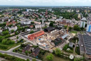 Widok z lotu ptaka na teren budowy Fabryki Pełnej Życia, z blokami mieszkalnymi, ulicami i zielenią w otoczeniu i w tle.