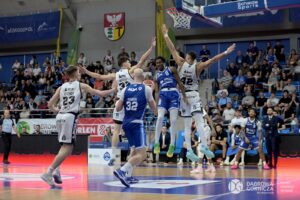 Dwóch koszykarzy z drużyny Legii, w jasnych strojach, próbuje zablokować rzut zawodnika MKS, w niebieskim stroju podczas meczu koszykówki przy pełnej trybunie hali sportowej.
