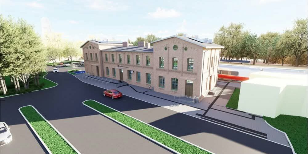 Widok z góry na kompleks dworca z cegły, parking i drogi dojazdowe; obok tory kolejowe z pociągiem.