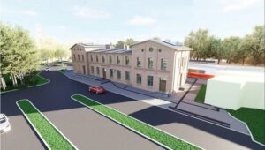 Widok z góry na kompleks dworca z cegły, parking i drogi dojazdowe; obok tory kolejowe z pociągiem.