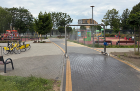 Skwer z infrastrukturą rowerową i placem zabaw, wyposażony w metalową konstrukcję z mgiełką wodną, służącą do chłodzenia.
