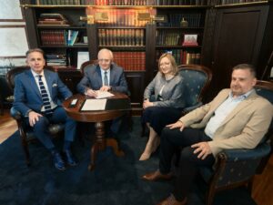 Cztery osoby siedzą przy okrągłym stoliku w eleganckim gabinecie z drewnianymi regałami wypełnionymi książkami; jedna osoba siedząca pośrodku podpisuje dokument, na stole leżą kartki i etui na długopis.