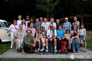 Grupa mieszkańców w różnym wieku pozuje do wspólnego zdjęcia przy nowo odsłoniętej ławce w parku. Ławka jest ciemna, z długim siedziskiem, stoi na jasnym mineralnym podłożu, wokół znajduje się trawnik. Uczestnicy siedzą i stoją wokół ławki, a w tle są drzewa oraz drewniana pergola. Po lewej stronie widoczny fragment przodu zabytkowego, jasnego Volkswagena T1.
