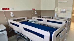 Nowoczesne łóżko szpitalne z granatowym materacem, z szafką przyłóżkową w sali wyposażonej w instalacje medyczne i umywalkę.