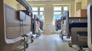 Rząd nowoczesnych łóżek szpitalnych z elektrycznym sterowaniem w przestronnej sali z dużymi oknami.