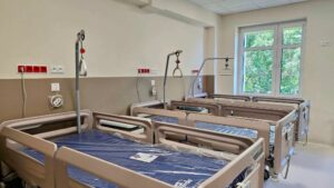 Jasna sala szpitalna wyposażona w kilka nowoczesnych łóżek rehabilitacyjnych z poręczami, uchwytami pomocniczymi i granatowymi materacami.