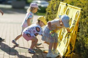 Troje dzieci w kolorowych ubraniach i czapeczkach na głowie bawi się przy żółtej planszy do gry zręcznościowej ustawionej na zewnątrz. Uczestnicy wrzucają woreczki do otworów oznaczonych punktami. Plansza stoi przy żywopłocie, a zabawa odbywa się na brukowanym chodniku w słoneczny dzień.