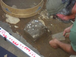 Archeolog prowadzi prace przy wykopie, w którym widoczne są srebrne monety odkryte w bryle ziemi. Na zdjęciu znajduje się także sprzęt pomiarowy i sito do przesiewania.