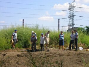 Uczestnicy badań archeologicznych stoją przy otwartym wykopie na terenie stanowiska archeologicznego. W tle widoczna jest roślinność, linie energetyczne oraz infrastruktura przemysłowa.