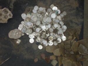 Zbiór srebrnych monet odkrytych podczas badań archeologicznych, widziane z góry na tle ciemnej ziemi i fragmentów naczyń ceramicznych.