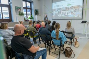 Uczestnicy szkolenia siedzą na krzesłach w sali konferencyjnej i słuchają prezentacji prowadzonej przed dużym ekranem. Na ekranie wyświetlona jest strona internetowa Dąbrowskiego Inkubatora Przedsiębiorczości. W pomieszczeniu o białych ścianach znajdują się duże okna, rośliny doniczkowe oraz stanowisko z laptopem do prezentacji.