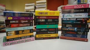 Alt text (wersja skrócona): Trzy stosy książek ustawione na stole w bibliotece. Na grzbietach widoczne są różne tytuły powieści popularnych polskich i zagranicznych autorów, a w tle znajduje się więcej książek.