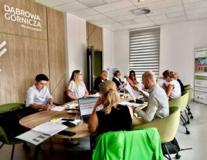 Spotkanie robocze przy stole konferencyjnym. Uczestnicy analizują dokumenty i prowadzą dyskusję, a w tle widoczne jest logo „Dąbrowa Górnicza dla aktywnych”.