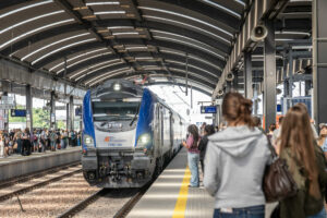 Niebiesko-szary pociąg Intercity wjeżdża na zadaszony peron dworca kolejowego. Po obu stronach torów stoją grupy podróżnych oczekujących na odjazd lub przyjazd pociągu. Widoczna jest charakterystyczna konstrukcja hali peronowej z łukowym dachem oraz oznaczenia peronów. Na czele składu znajduje się lokomotywa EU160-094 z logo PKP Intercity.