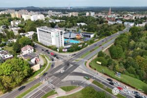 Duże skrzyżowanie ze światłami. Z prawej strony drzewa i krzewy, w górnej części biały budynek hotelu, a obok aquapark.