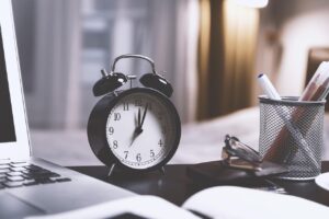 Czarny budzik na biurku obok laptopa, okularów i pojemnika z długopisami. W tle rozmyte wnętrze z ciepłym światłem.