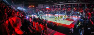 Hala sportowa podczas meczu koszykówki. Na oświetlonym parkiecie stoją dwie drużyny ustawione przed rozpoczęciem spotkania. Trybuny wypełniają kibice, a nad boiskiem widoczne są efekty świetlne i telebimy tworzące oprawę wydarzenia.