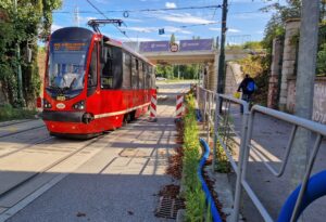 Czerwony tramwaj linii 27 jedzie po torowisku, w tle wiadukt. Po prawej stronie znajduje się chodnik z metalowymi barierkami oraz osoba idąca w stronę przejścia pod wiaduktem. Słoneczny dzień, bezchmurne niebo z nielicznymi chmurami.